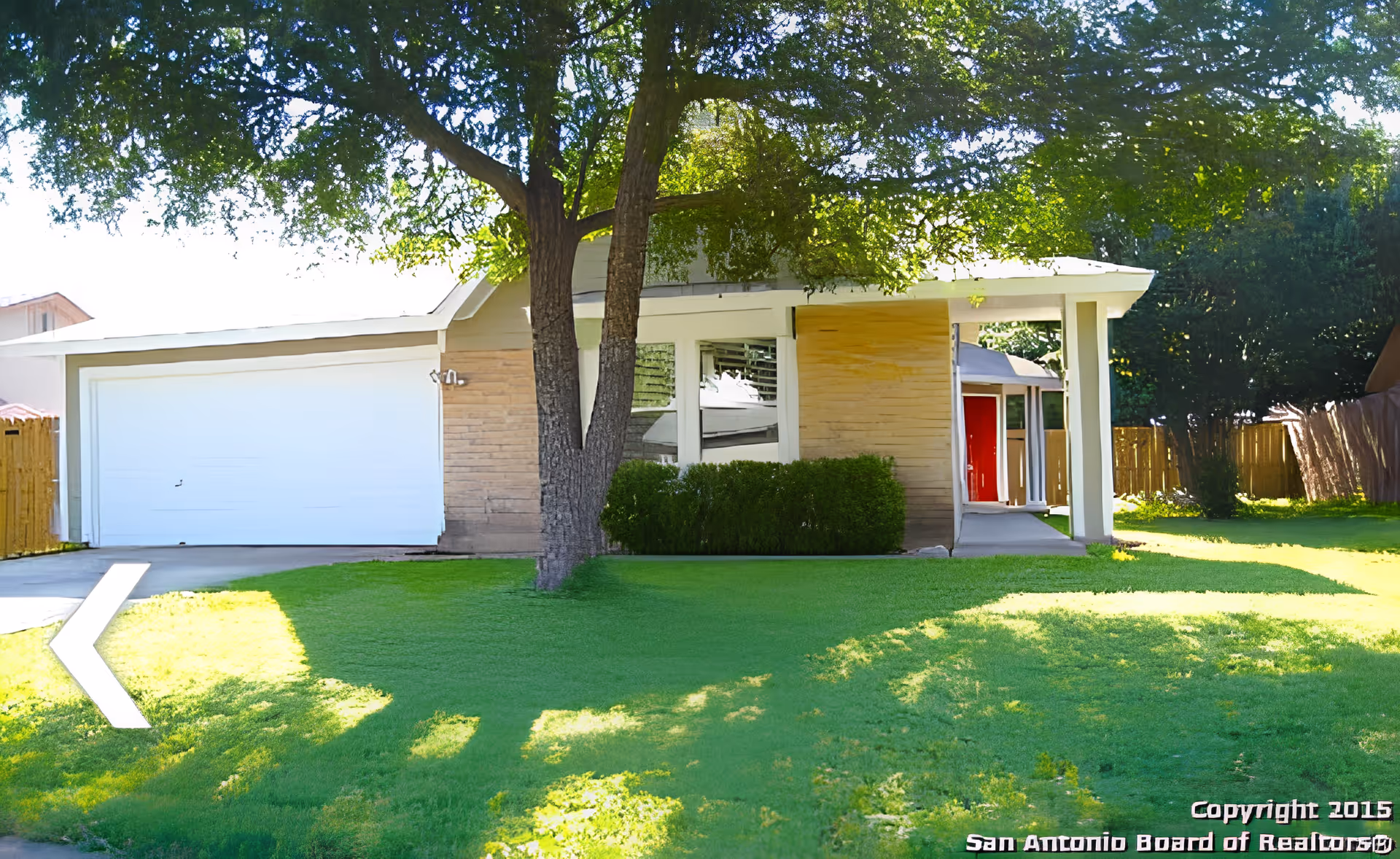 Exterior view of a single-story residential building with a large tree in the front yard, green grass, a white garage door, and a red front door under a covered porch.