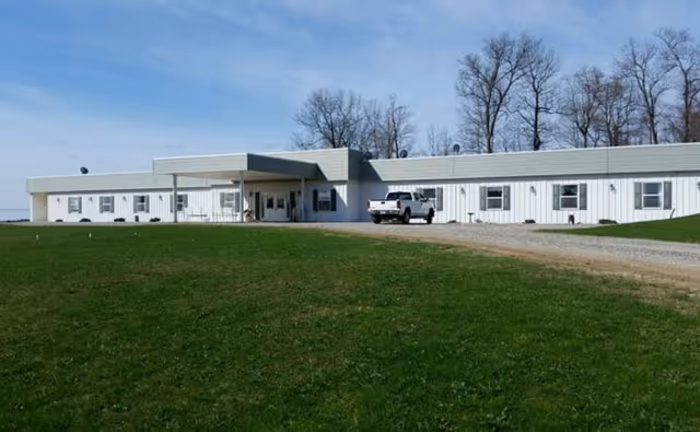 Single-story white building with a covered entrance and several windows, situated on a grassy area with a gravel driveway and a white pickup truck parked near the entrance. Leafless trees are visible in the background under a clear blue sky.