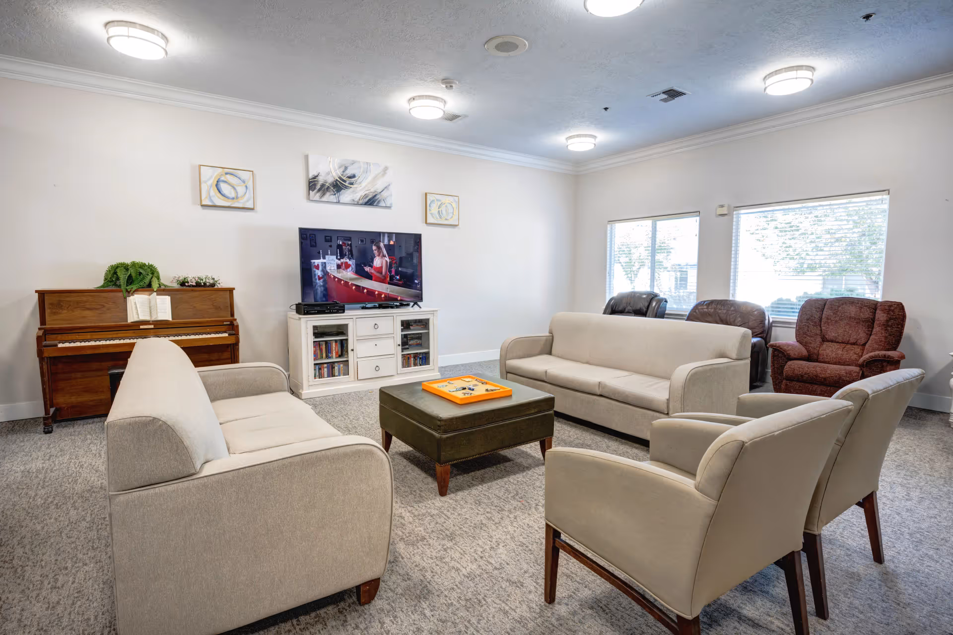 A senior living facility common room with two beige sofas, two beige armchairs, a dark green ottoman with a board game on top, a wooden piano with a plant and sheet music, a white TV stand with a flat-screen TV showing a woman in a red dress, and three windows letting in natural light.