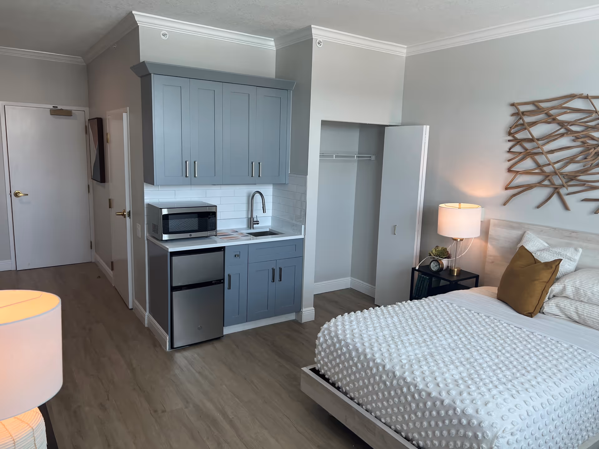 A studio apartment room featuring a bed with white textured bedding and a brown pillow, a bedside table with a lamp and clock, a small kitchenette with blue cabinets, a microwave, a mini fridge, and a sink. There is an open closet space next to the kitchenette and a closed door in the background. The room has light wood flooring and neutral-colored walls.