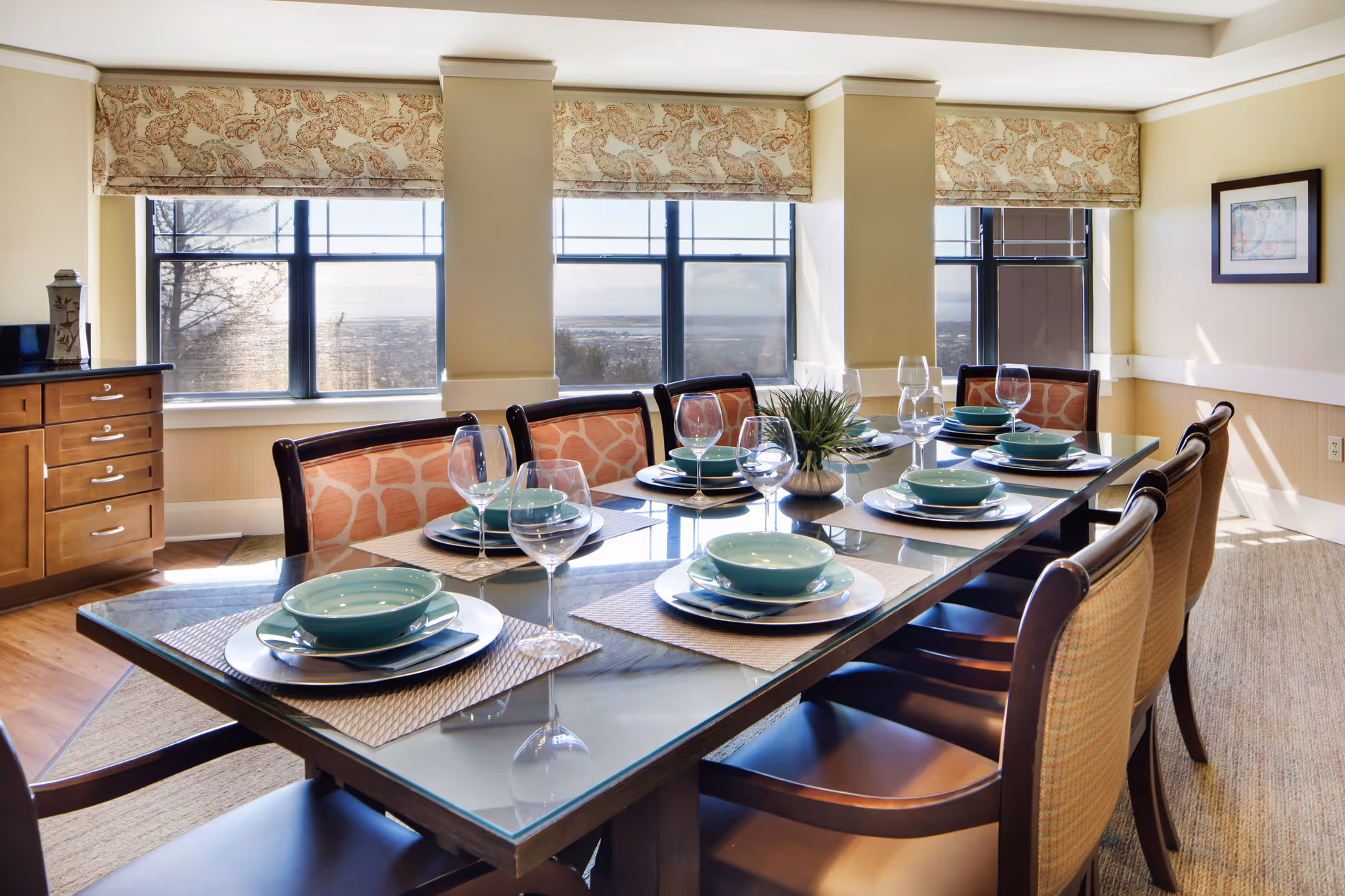 A bright dining room with a large glass-top table set for eight people. Each place setting includes a turquoise bowl, plate, napkin, and wine glass. The chairs have wooden frames with patterned upholstery. Large windows with patterned valances let in natural light and offer a view outside. A small plant centerpiece is on the table, and wooden cabinetry is visible on the left side of the room.