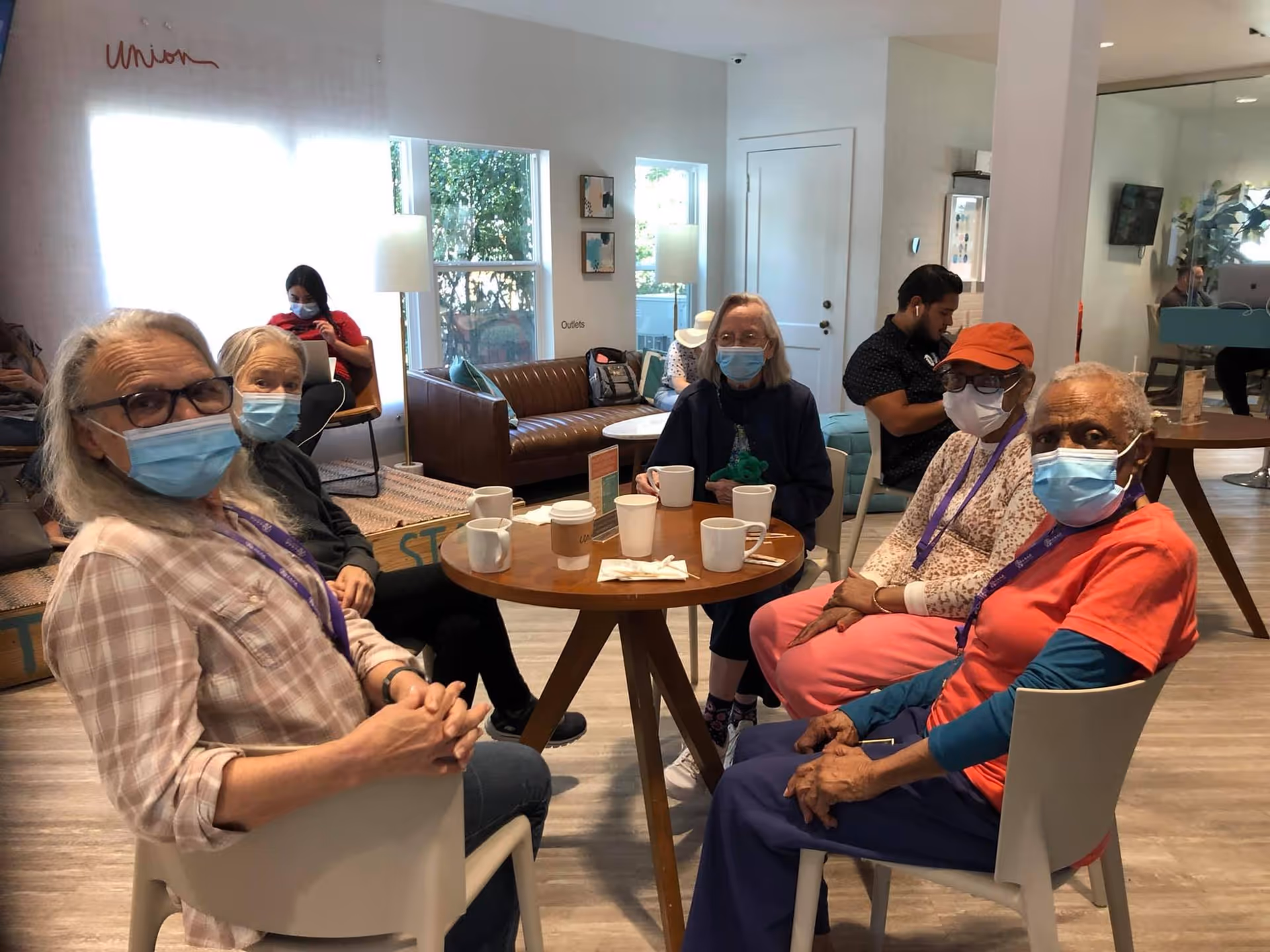 A group of elderly individuals wearing face masks sitting around a round wooden table with several coffee cups in a well-lit common area. The room has large windows, modern furniture including a brown leather couch, and light-colored walls. A few other people are visible in the background, some seated and engaged with electronic devices.
