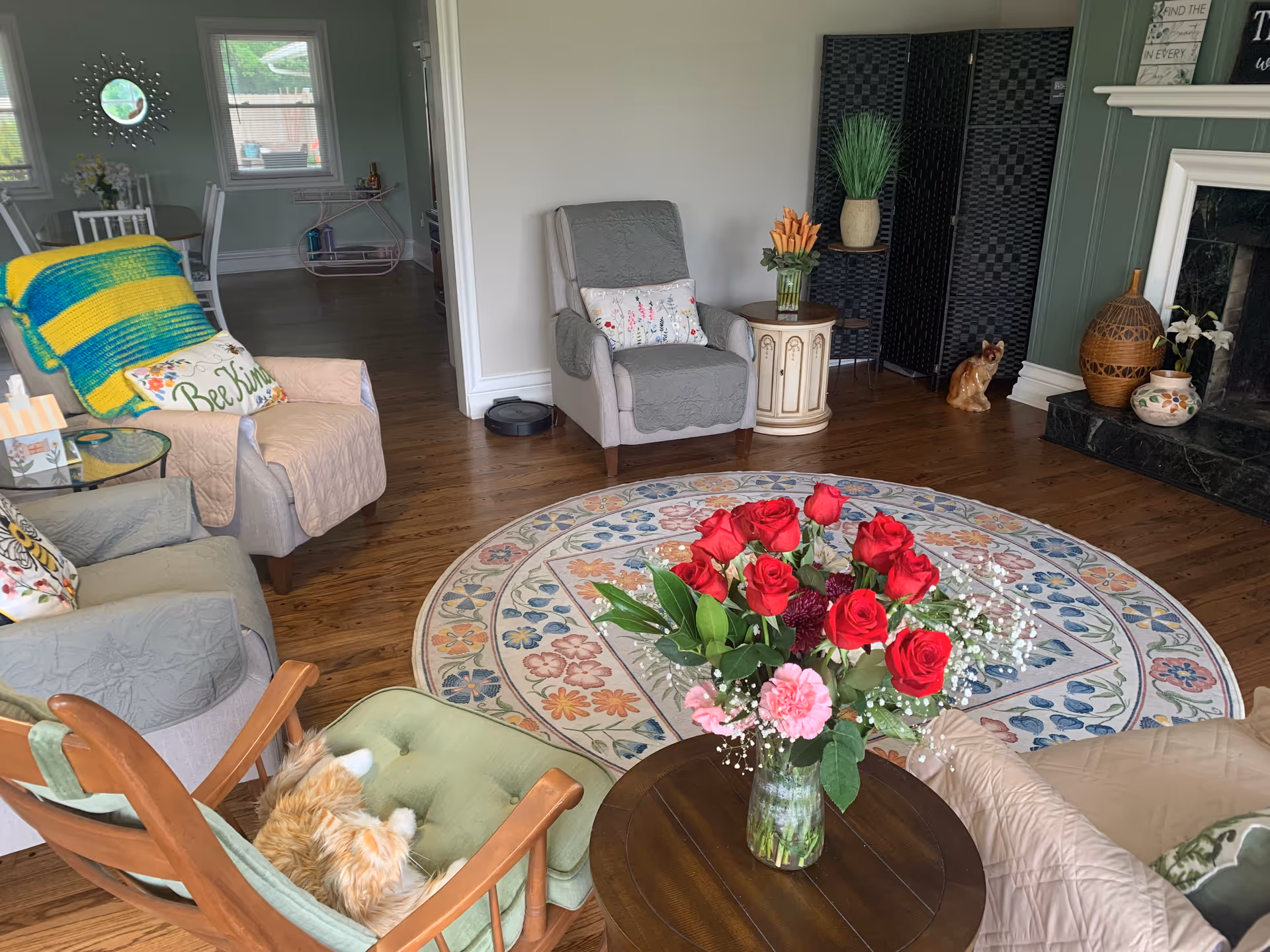 A cozy living room with several armchairs and a rocking chair arranged around a round floral-patterned rug. A vase with red and pink flowers sits on a wooden table in the center. The room has wooden flooring, a fireplace with decorative vases, and a green accent wall. There is a window and a doorway leading to another room with a dining table and chairs.