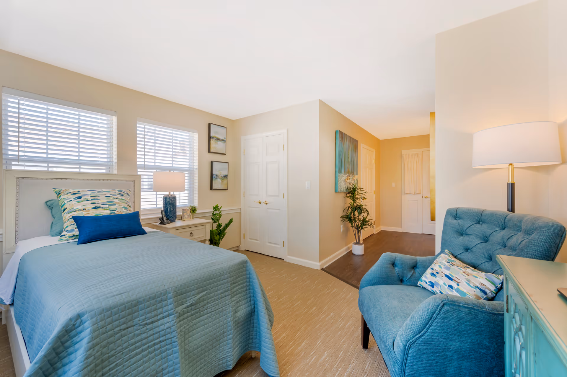 A bright and cozy bedroom with a single bed covered in a light blue quilt and decorative pillows. Next to the bed is a white nightstand with a blue lamp and a small plant. Two windows with white blinds let in natural light. A comfortable blue armchair with a patterned pillow is positioned near a floor lamp and a turquoise cabinet. The room has beige walls and carpet, with a hallway leading to white doors and a wooden floor area.