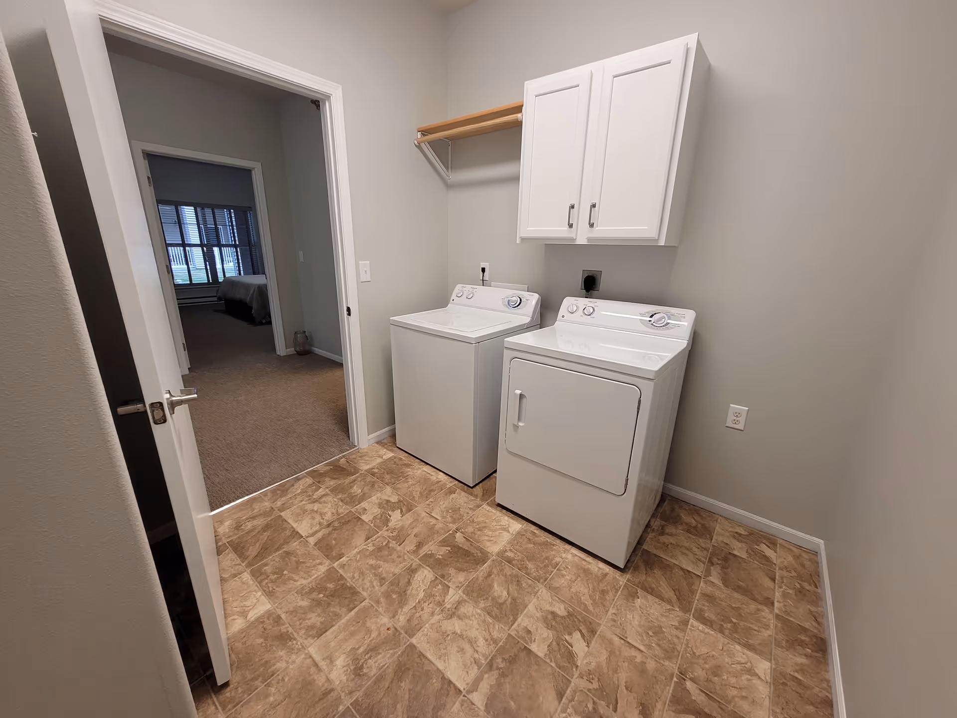 Laundry room with a white washing machine and dryer side by side, a white cabinet mounted on the wall above the dryer, and a wooden shelf with a hanging rod above the washing machine. The floor has brown tile, and there is an open door leading to a carpeted bedroom with a bed and large windows.