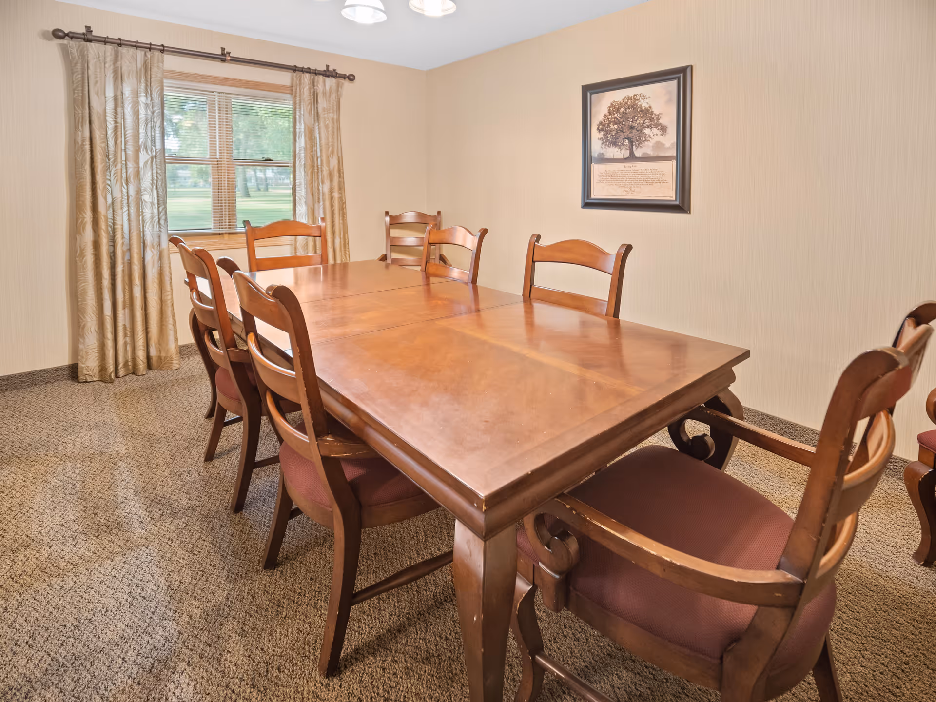 A dining room with a large wooden table surrounded by eight wooden chairs with padded seats. There is a window with beige patterned curtains on one wall and a framed picture of a tree on another wall. The room has beige walls and carpeted flooring.