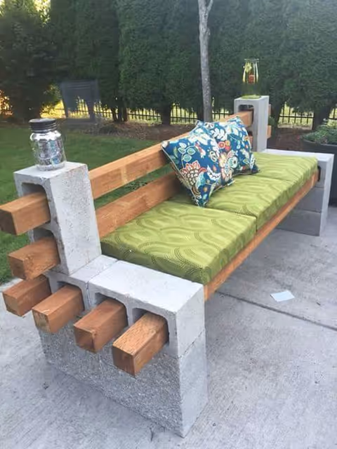 Outdoor bench made with concrete blocks and wooden beams, featuring a green cushion and a colorful floral pillow, placed on a concrete patio with greenery and a fence in the background.
