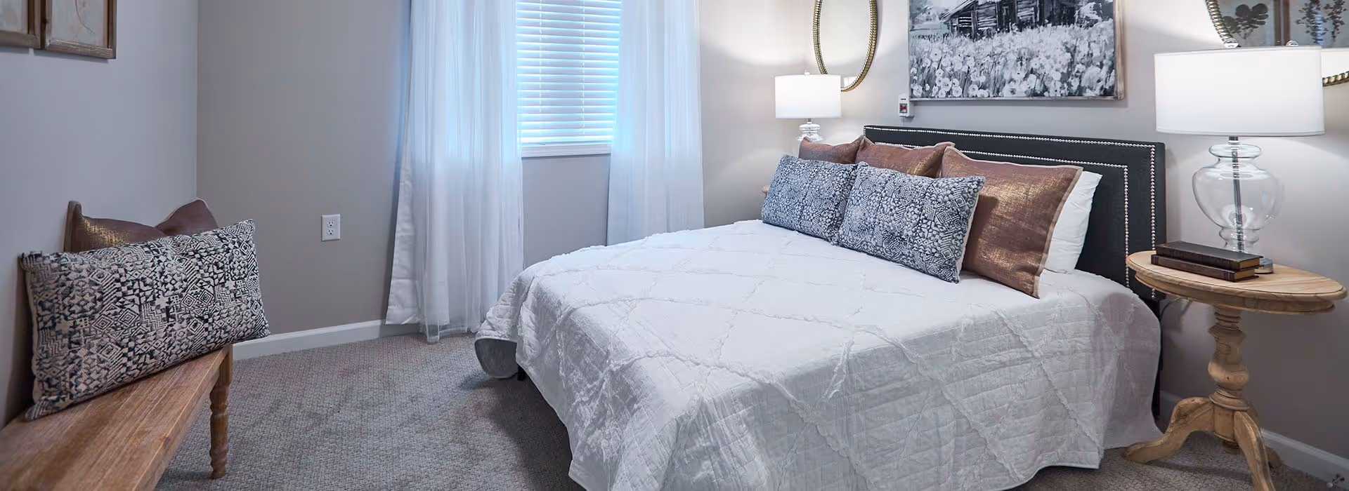 A cozy bedroom with a neatly made bed featuring a white quilt and multiple decorative pillows. There is a wooden bench with patterned cushions on the left side, a window with white curtains, a round wooden side table with a glass lamp and books, and a wall mirror and artwork above the bed.