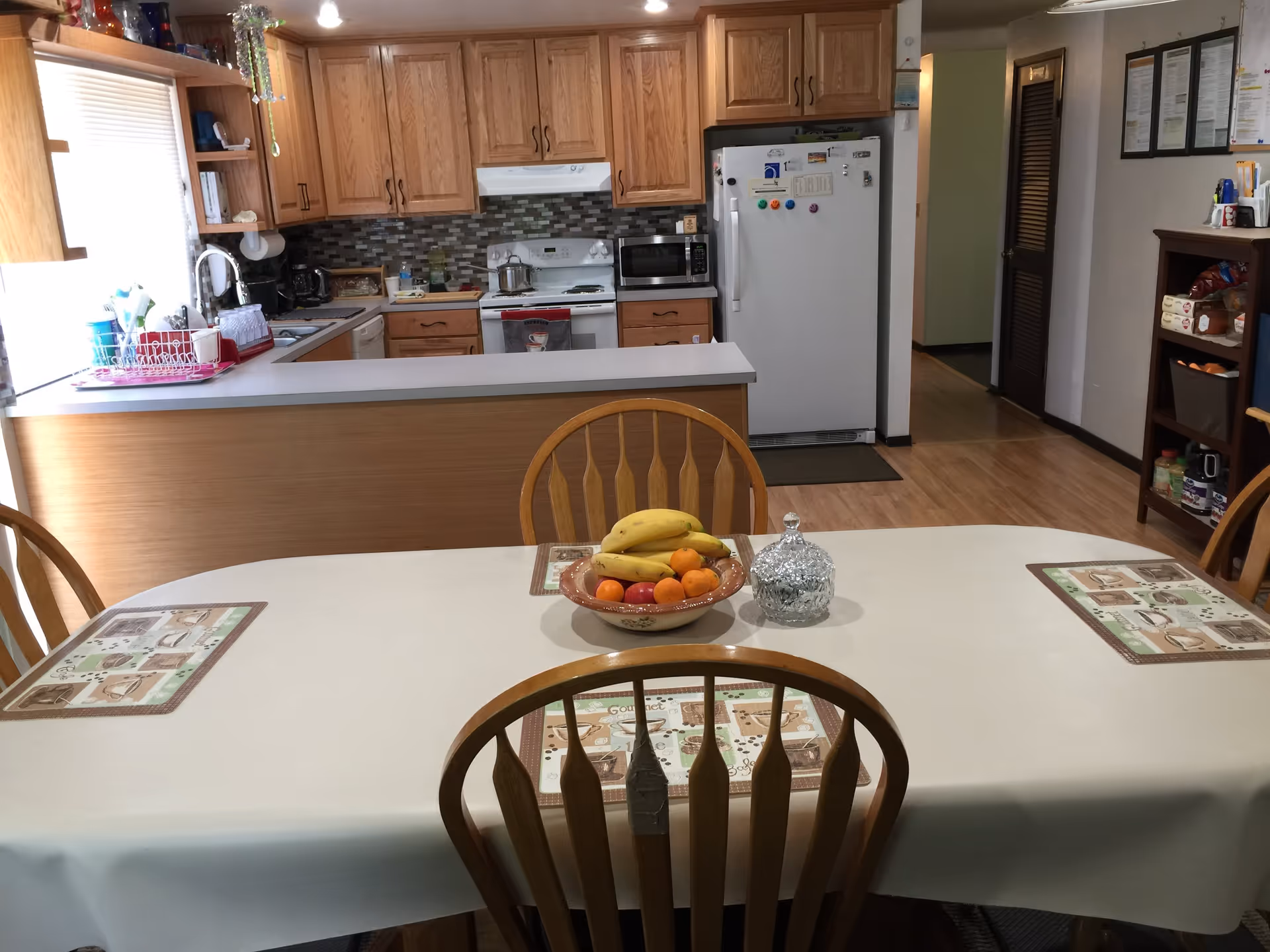 A kitchen and dining area with wooden cabinets, a white refrigerator, stove, microwave, and a sink. A dining table covered with a white tablecloth has four wooden chairs around it and a bowl of fruit including bananas and oranges in the center. The floor is wooden, and there is a small shelf with snacks and other items on the right side.