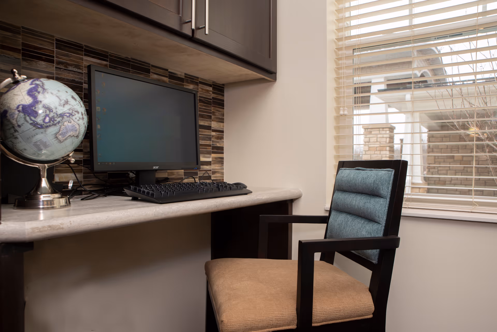 A small workspace with a computer monitor and keyboard on a marble countertop. A globe is placed to the left of the monitor. There is a cushioned chair with a wooden frame in front of the desk. A window with blinds is on the right side, showing part of a building exterior.