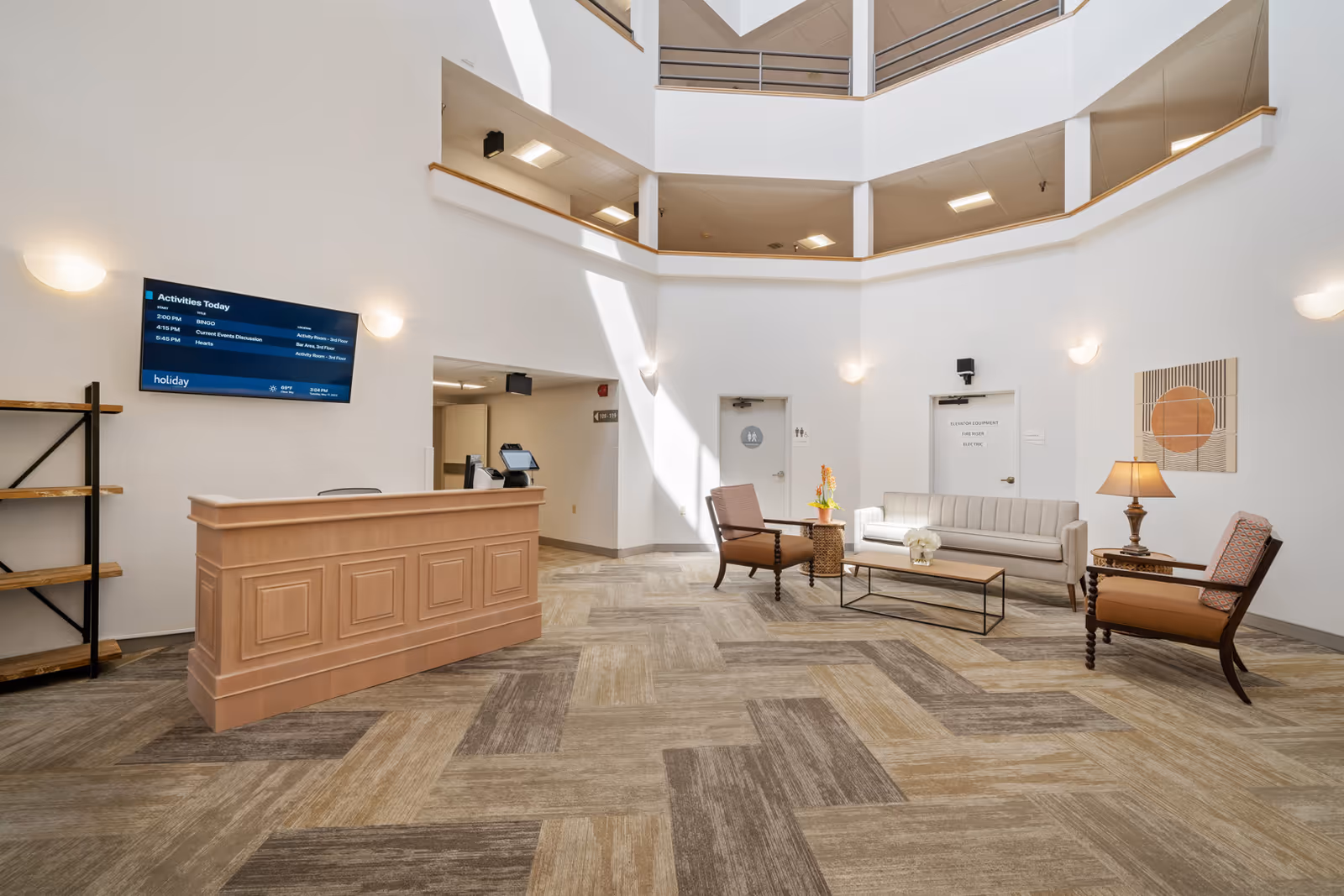 Bright, modern atrium lobby with a reception desk, seating area of sofa and chairs, and a wall-mounted TV.