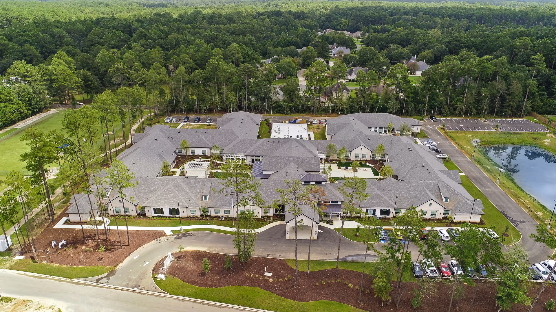 Aerial view of a large senior living facility named Sage Lake surrounded by trees and greenery. The building has multiple wings with gray roofs and white walls, parking lots with cars, and landscaped grounds including a pond on the right side.