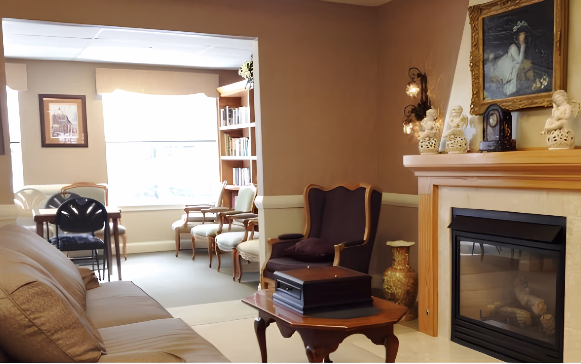 Cozy senior living room with sofa, armchairs, a fireplace, coffee table and a bookshelf by a large window.