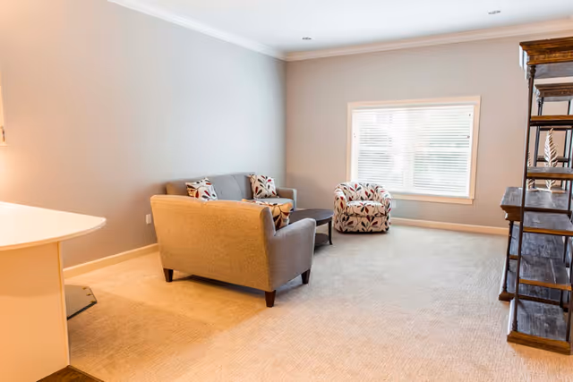 A bright living room with beige carpet and light gray walls. The room features a gray loveseat with patterned cushions, a matching patterned armchair, a round dark coffee table, and a wooden shelving unit on the right. A large window with white blinds allows natural light to fill the space.