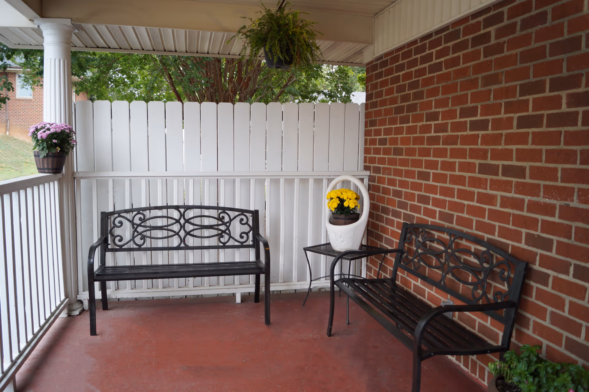 Covered porch with two decorative metal benches facing a white fence, a brick wall, and potted flowers.