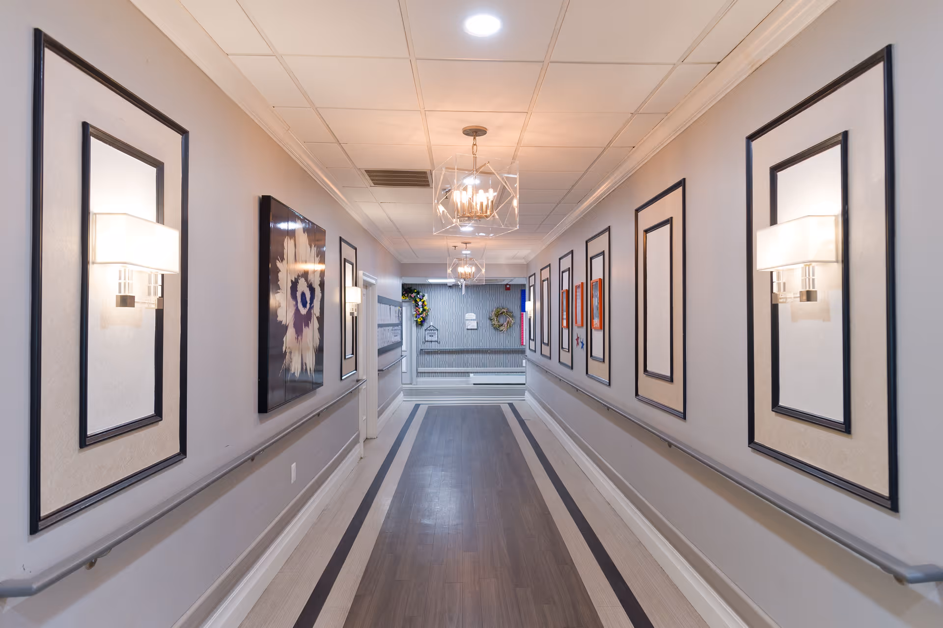 A well-lit hallway in a senior living facility with light gray walls decorated with framed artwork and wall sconces. The floor has a wood-like finish with a patterned border, and there are handrails on both sides of the hallway. At the end of the hallway, there are wreaths and a bulletin board on the wall.