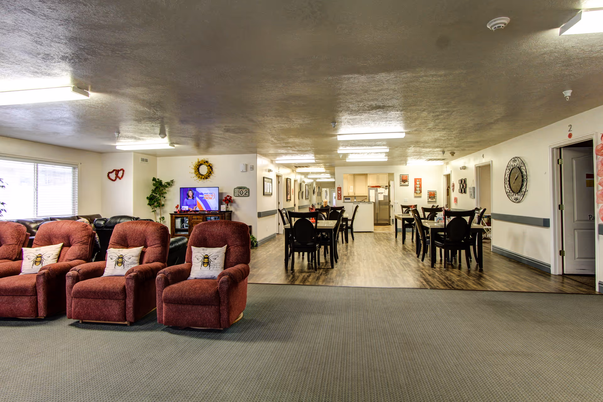 Interior view of a senior living facility common area featuring a seating area with four red recliners each with a pillow decorated with a bee design, a television on a stand, several dining tables with chairs, and a kitchen area in the background. The room has a carpeted floor in the seating area and wood flooring in the dining and kitchen area, with bright overhead lighting and wall decorations including a clock and framed pictures.
