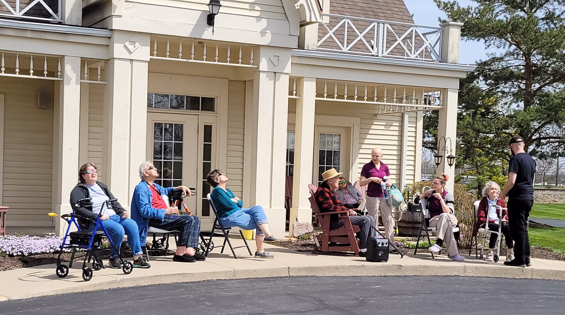 A group of elderly people and caregivers sitting and standing outside a beige building with white trim. Some individuals are seated in wheelchairs and chairs, enjoying the sunny day on a paved area near the entrance. There are trees and grass in the background.