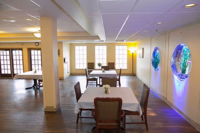 Bright communal dining room with tables draped in white linens, wooden chairs, large windows, and illuminated wall aquariums.