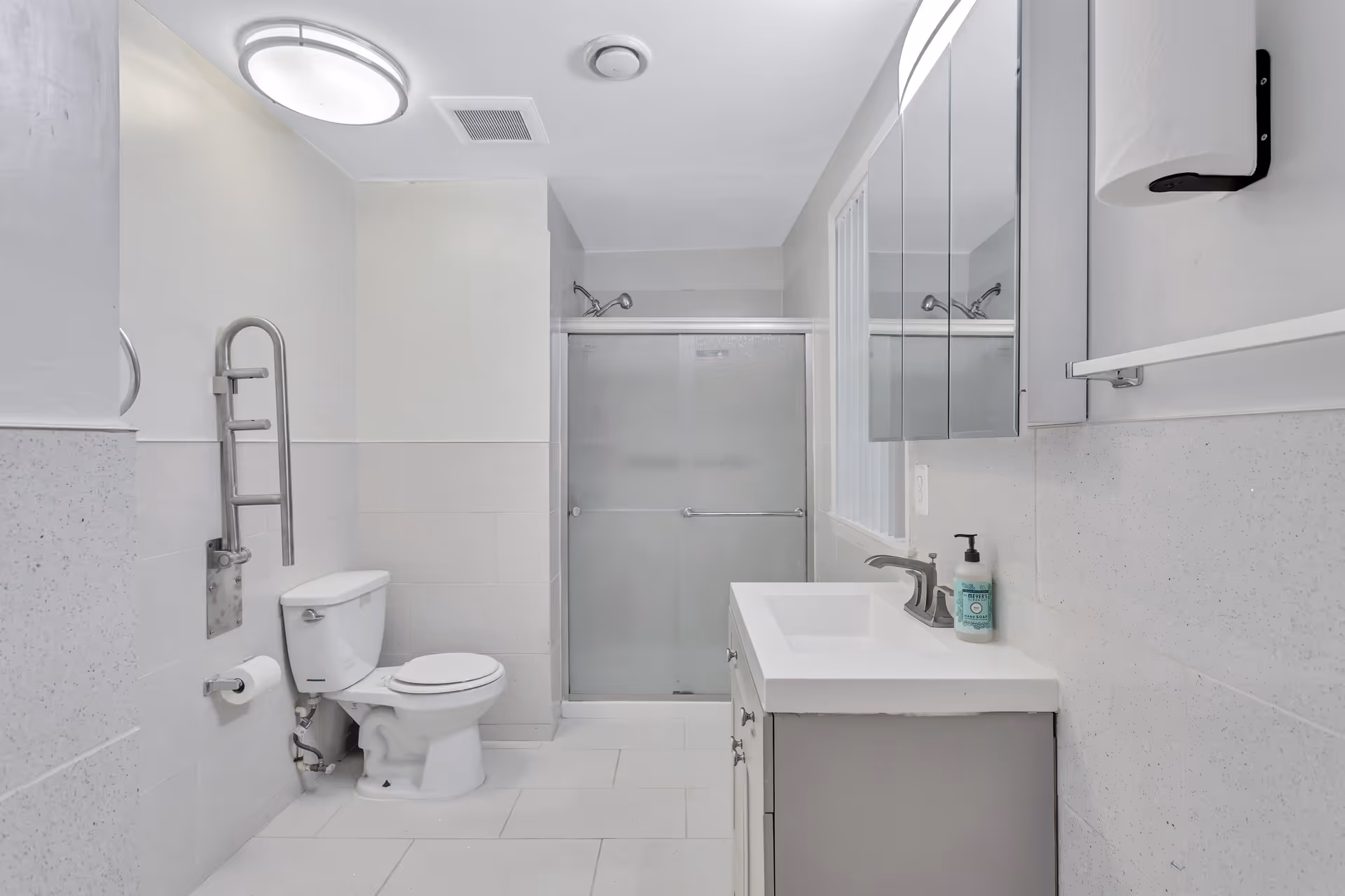 A clean, modern bathroom featuring a toilet with a stainless steel grab bar, a sink with a faucet and a bottle of hand soap, a mirrored medicine cabinet above the sink, a paper towel dispenser on the wall, and a shower with frosted sliding glass doors.