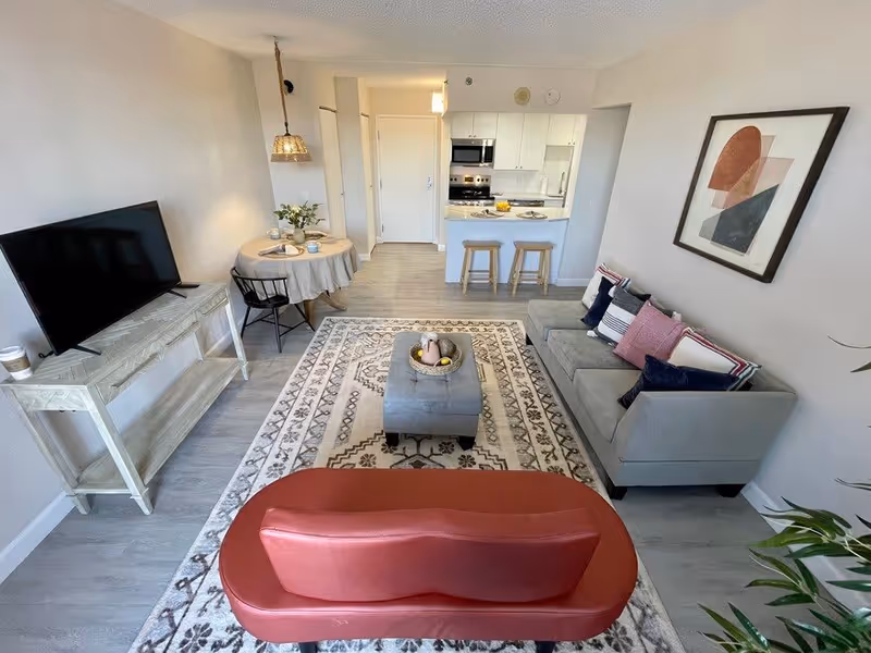 A modern living room and kitchen area with a gray sofa adorned with colorful pillows, a patterned area rug, a red bench, a small round dining table with two chairs, a flat-screen TV on a wooden stand, and a kitchen with white cabinets and two wooden bar stools.