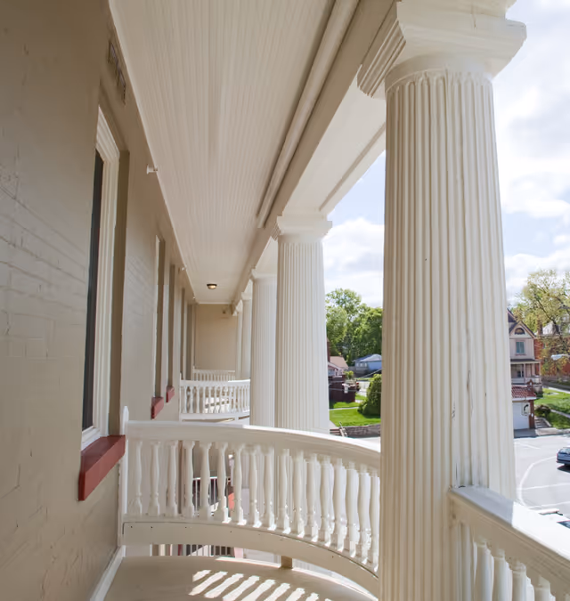 View of a balcony with white balustrades and large white columns, overlooking a parking area and some houses with trees in the background under a partly cloudy sky.