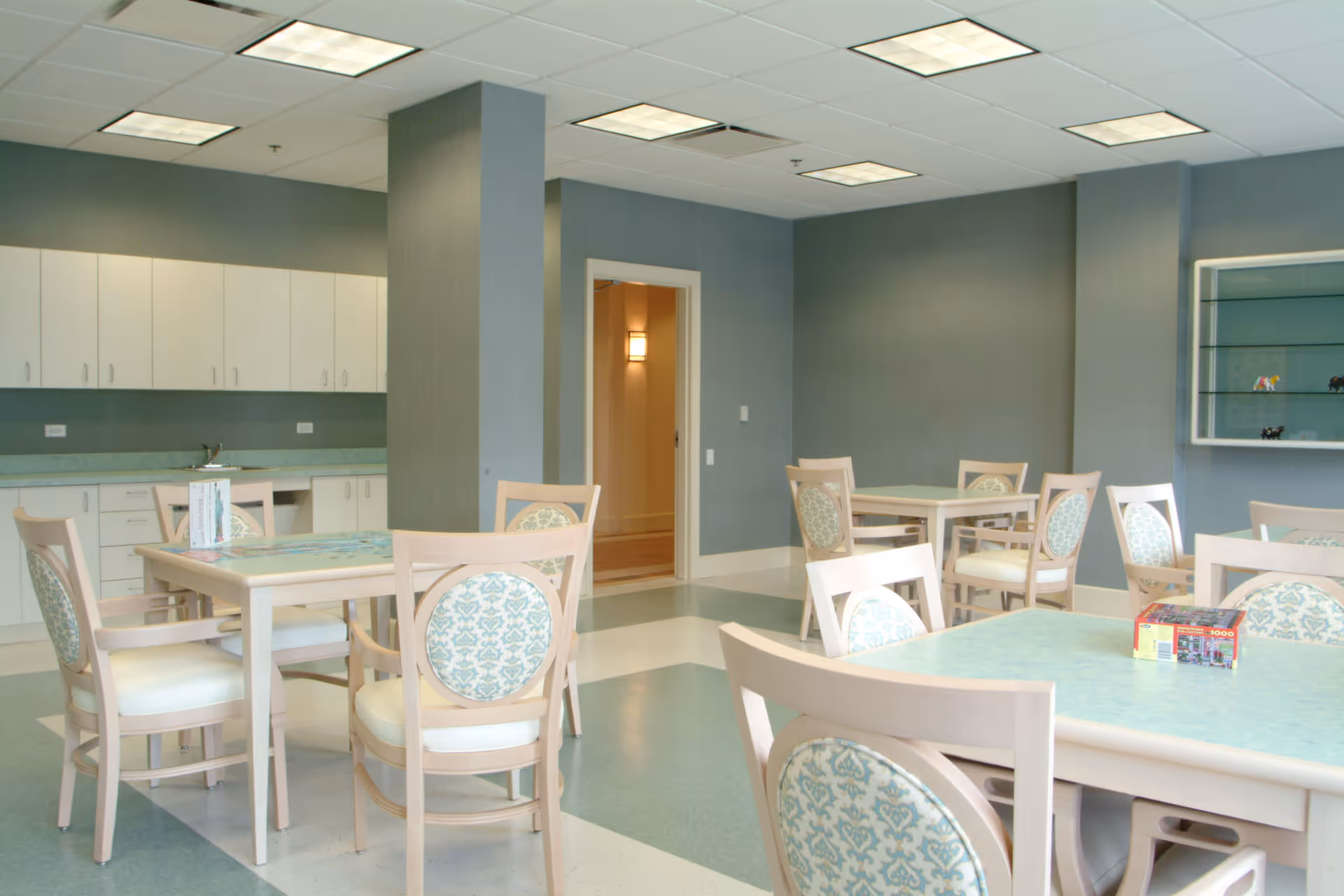 A bright and clean dining room with several tables and chairs. The chairs have light wood frames and patterned upholstery. The room features blue walls, a tiled floor with a blue and white pattern, and white cabinets along one wall. There is a doorway leading to another room and a glass display case on the right wall.