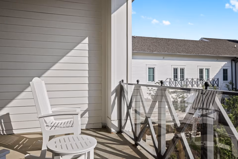 Sunlit balcony with white chairs and a small round table beside a glass railing overlooking a neighboring building.