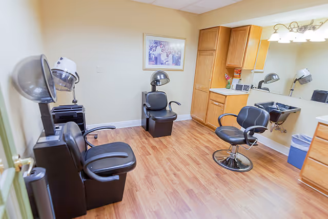 Interior view of a salon area in a senior living facility with three black salon chairs, two hair dryers, wooden cabinets, a large mirror, and a framed picture on the wall. The floor is wood laminate and the room is well-lit.