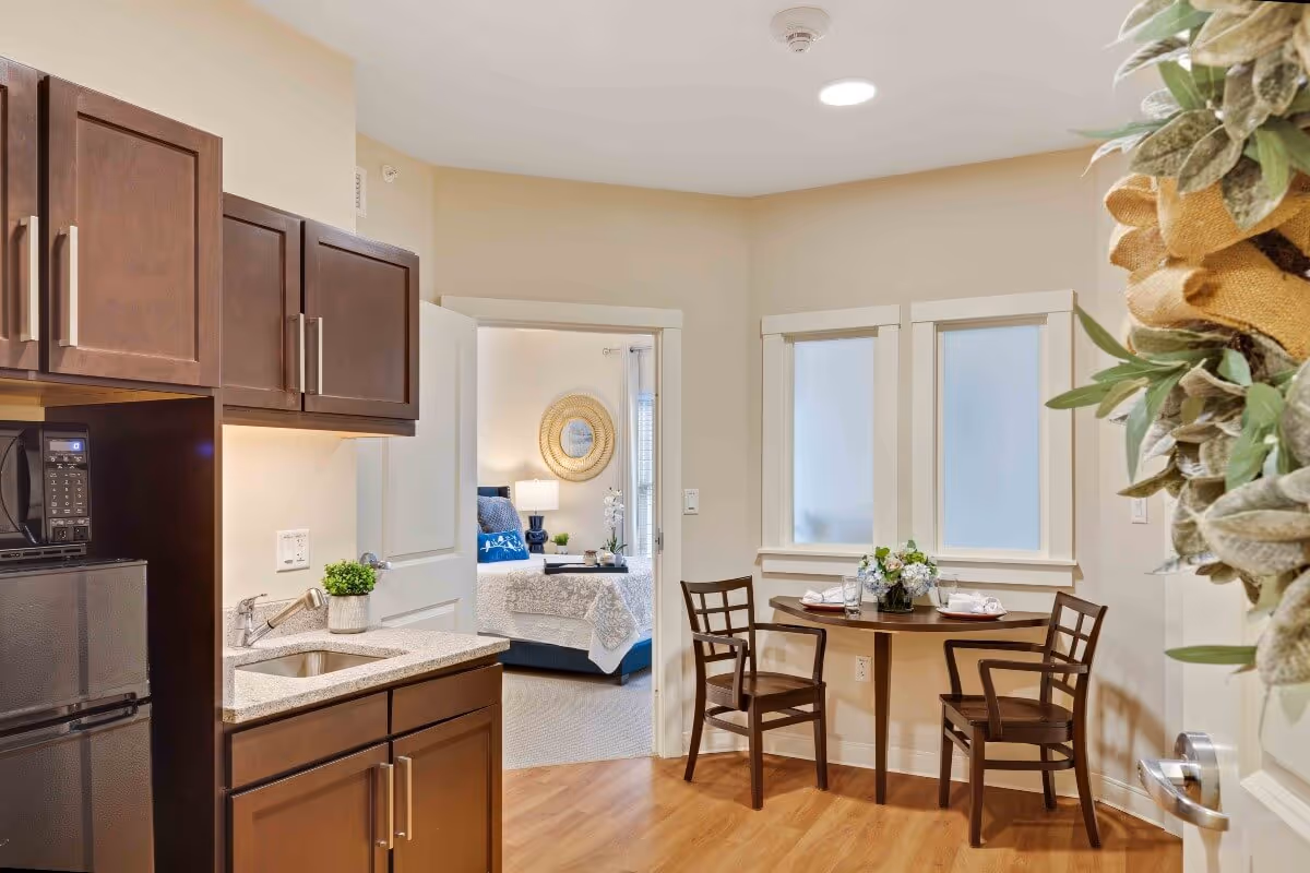 Small apartment interior showing a kitchenette with dark cabinets, a round dining table with two chairs, and a bedroom visible through an open door.
