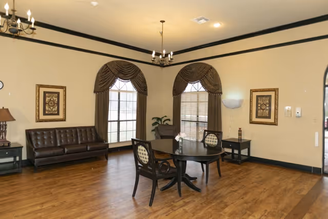 A spacious room with wooden flooring featuring a dark brown leather sofa against the wall, two large windows with brown drapes, a round black table with two chairs, two framed artworks on the walls, a floor lamp, and a side table with a lamp. The room has beige walls with dark trim and two chandeliers hanging from the ceiling.