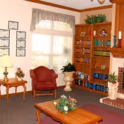 Cozy living room with a red armchair, wooden coffee and side tables, a bookshelf, fireplace, and a large window with curtains.