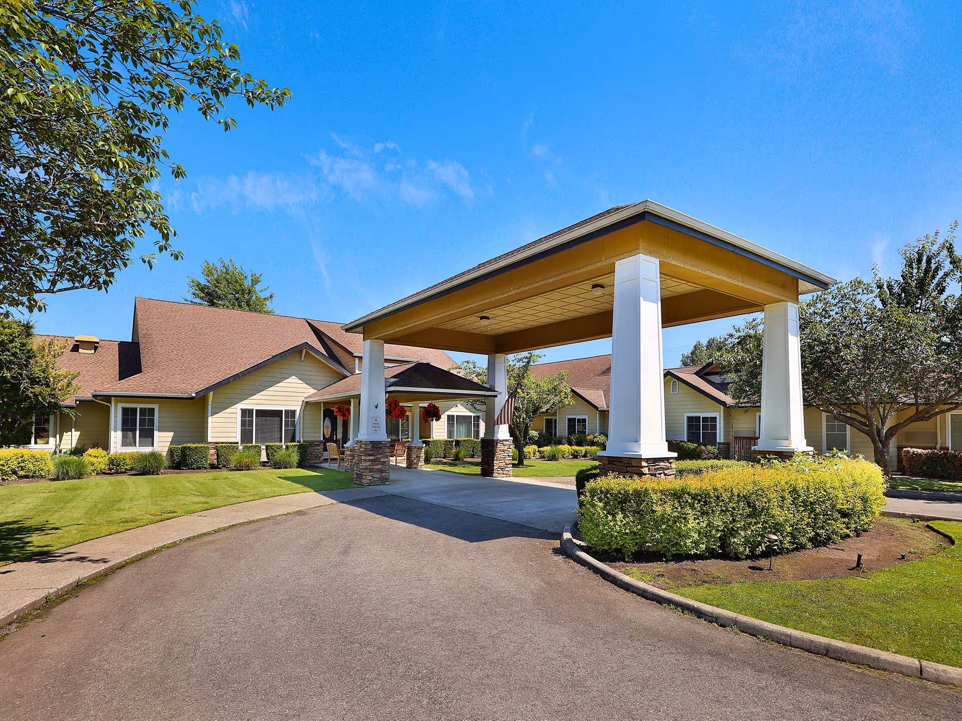 Exterior view of White River Memory Care facility showing a covered entrance with four large white pillars and a driveway leading up to it. The building has beige siding with brown roofs, surrounded by green lawns, bushes, and trees under a clear blue sky.
