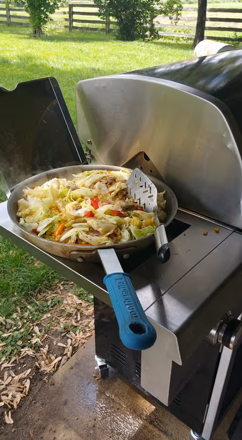 A skillet of stir-fried vegetables sits on the side shelf of a gas grill with a grassy yard and wooden fence in the background.