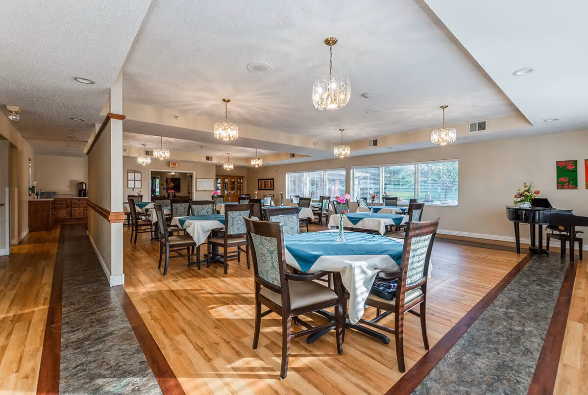 Bright communal dining room with multiple tables draped in teal tablecloths, chairs, chandeliers, and a piano near large windows.