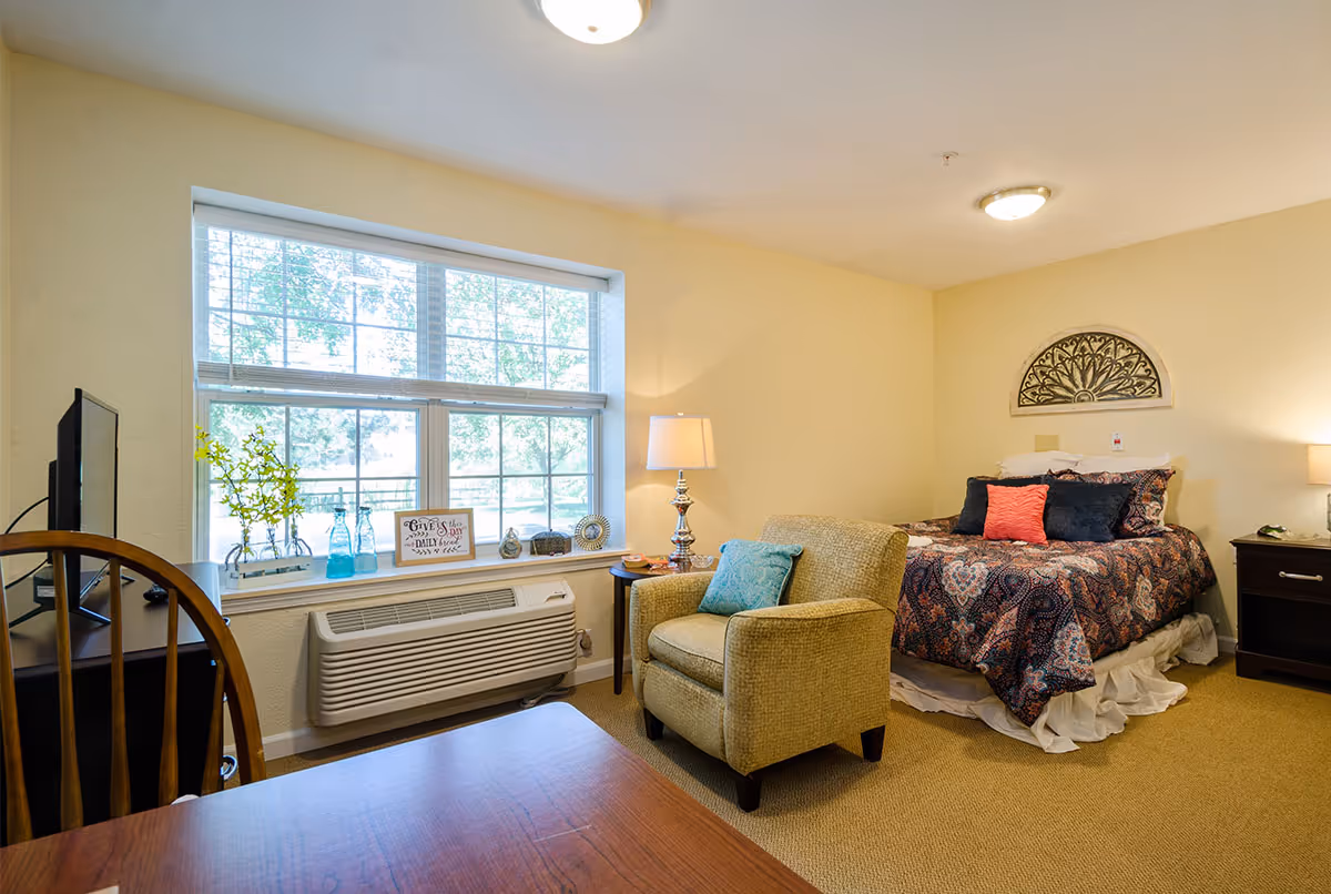 Well-lit studio bedroom with a patterned bed, upholstered armchair, desk, and large window overlooking greenery.
