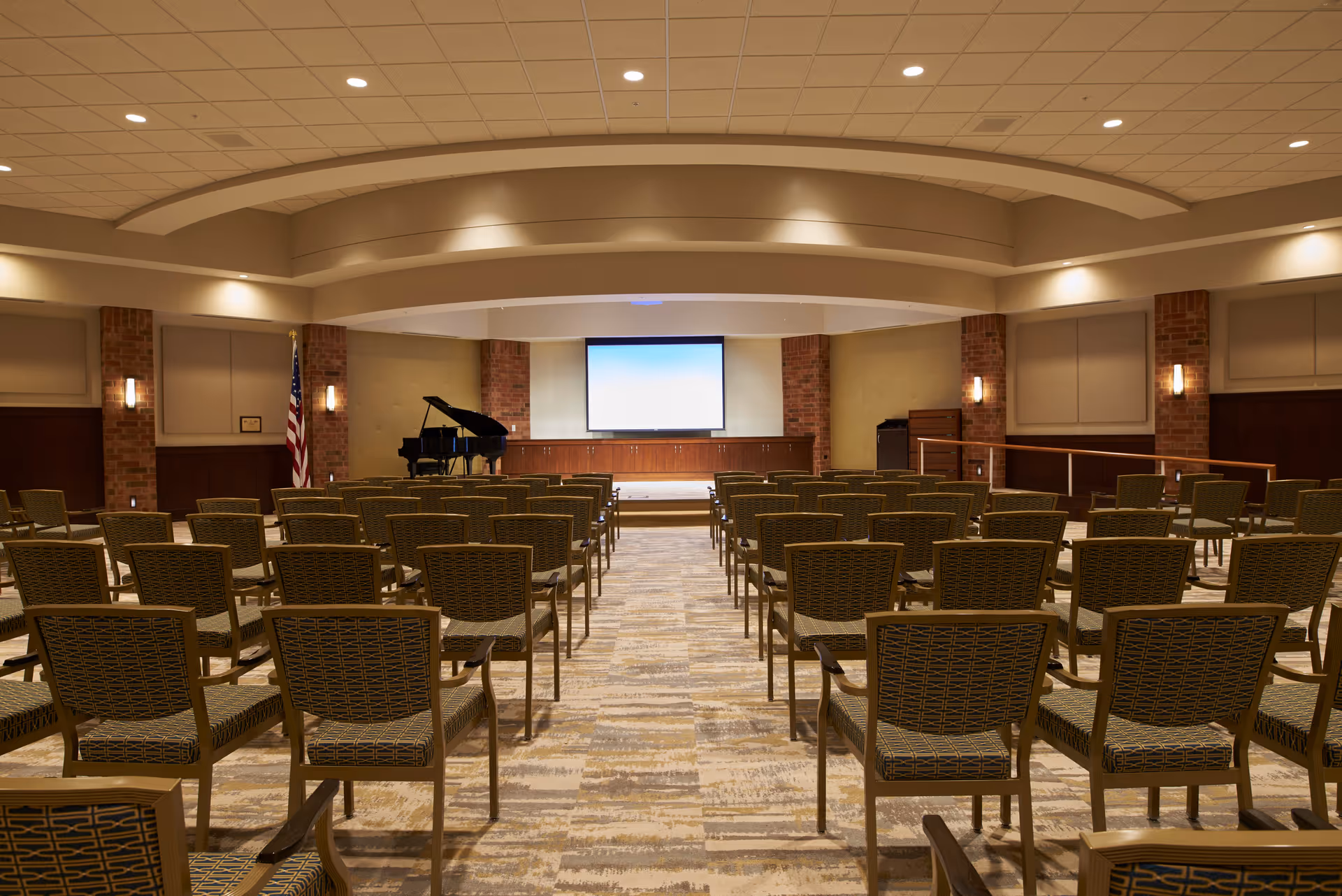 Spacious auditorium-style meeting room with rows of chairs facing a stage with a piano and projection screen.