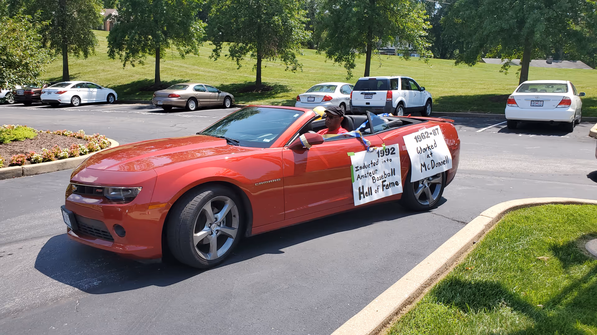 A red convertible Chevrolet Camaro with two signs attached to the passenger side door. One sign reads '1992 Inducted into Amateur Baseball Hall of Fame' and the other reads '1962-87 Worked at McDonnell'. A person wearing a hat is sitting in the driver's seat. The car is parked on a paved area with grass and trees in the background.
