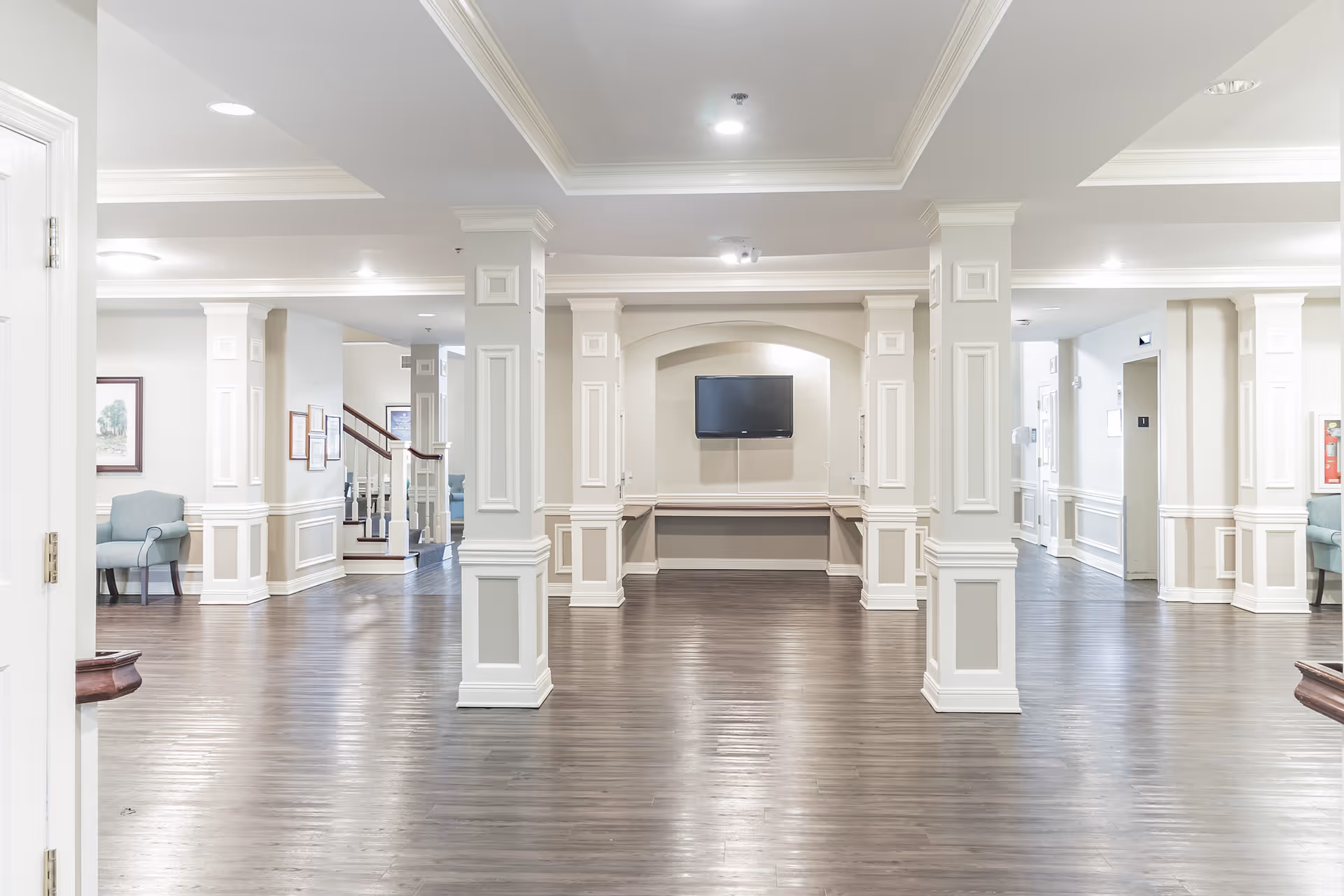 Spacious bright common area with decorative columns, hardwood floors, seating and a wall-mounted TV.