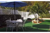 Shaded outdoor patio with a white metal table and chairs under a blue umbrella, artificial turf, potted palms and a white fence.