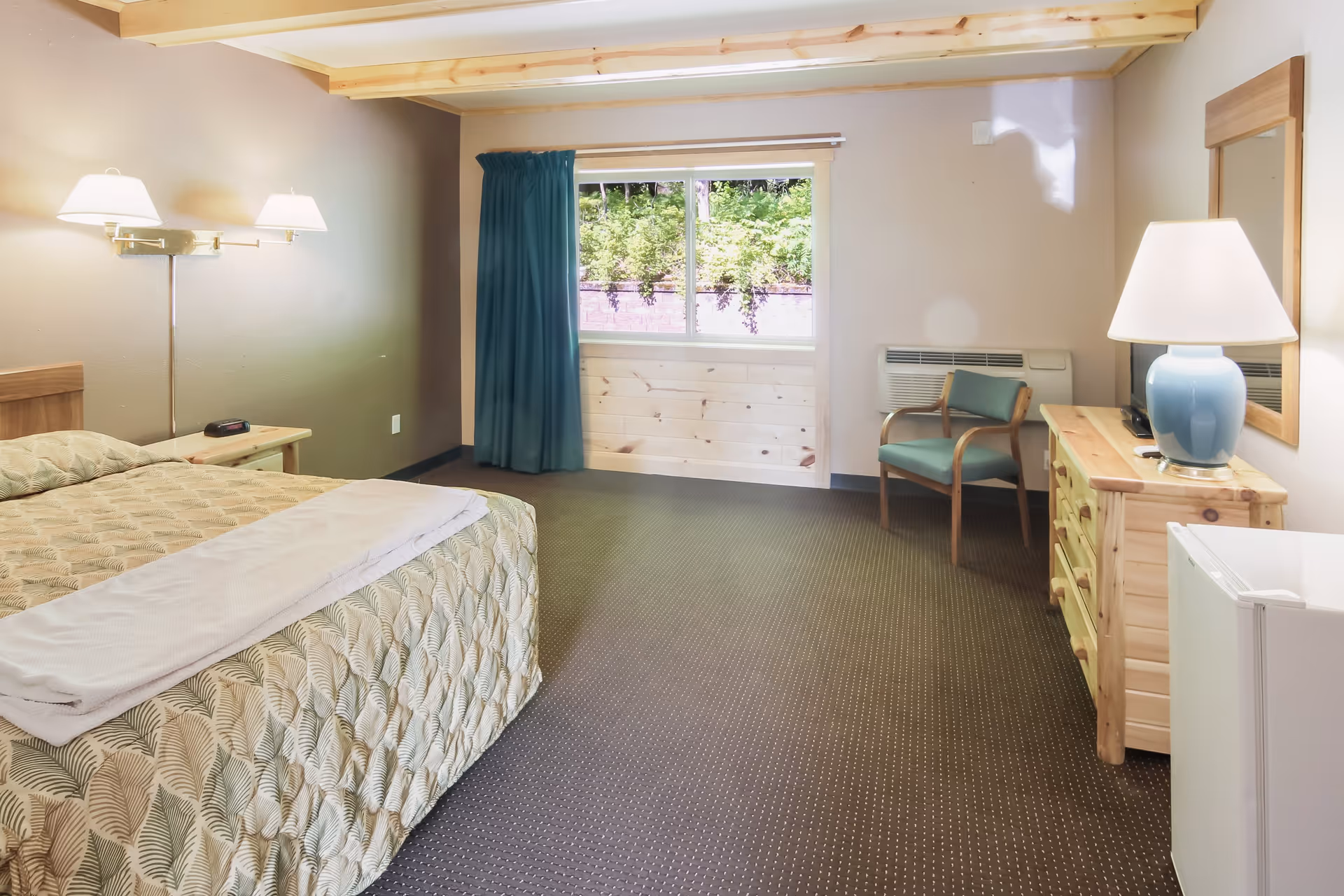 A cozy bedroom in South Torch Assisted Living featuring a bed with patterned bedding and a folded white blanket on top, a wooden nightstand with a clock, a wall-mounted double lamp, a window with blue curtains showing greenery outside, a wooden dresser with a blue lamp and mirror above it, a green cushioned chair, and a small white refrigerator.