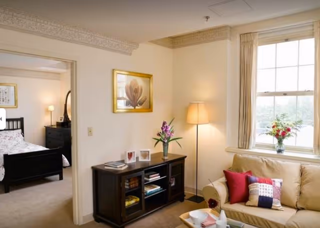 A cozy living room with a beige sofa adorned with colorful pillows next to a window with beige curtains. A floor lamp stands in the corner, and a dark wooden cabinet with books and decorative items is against the wall. A framed floral picture hangs above the cabinet. Through an open doorway, a bedroom with a black bed frame and a bedside lamp is visible.