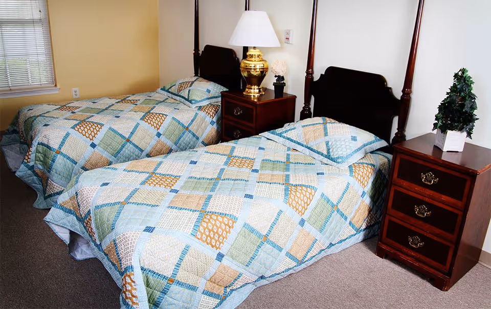 A bedroom with two twin beds featuring matching patchwork quilts in shades of blue, green, and beige. Between the beds is a wooden nightstand with a brass lamp and a small decorative item. Another wooden nightstand with a small potted plant is beside the bed on the right. The room has beige walls and a window with blinds on the left side.