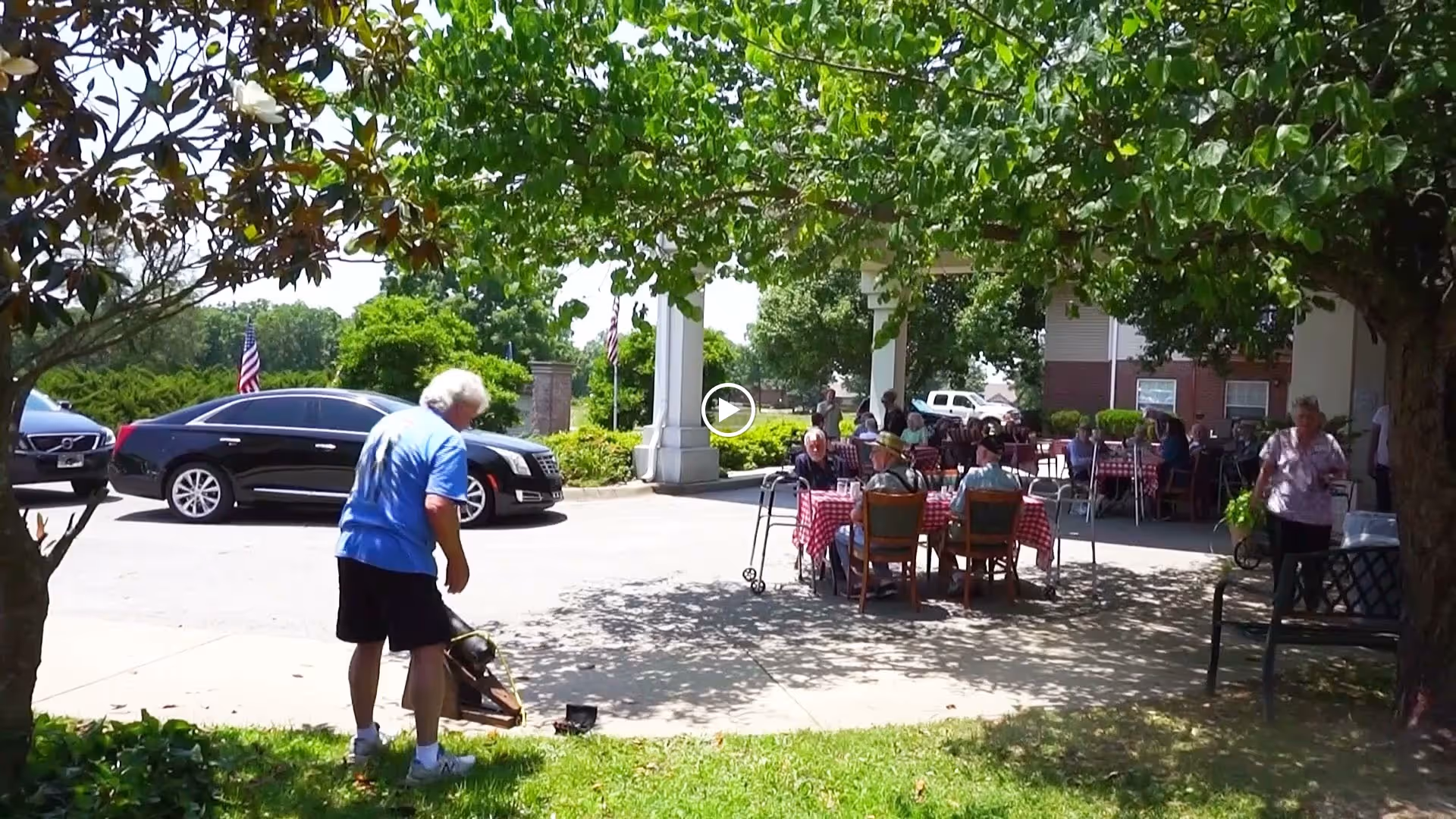 Outdoor patio area at Peachtree Village Assisted Living Holiday Island with senior residents sitting at tables covered with red and white checkered tablecloths under a shaded area. A man in a blue shirt and black shorts is standing near a tree in the foreground, and several cars are parked nearby. Trees provide shade and greenery around the space.