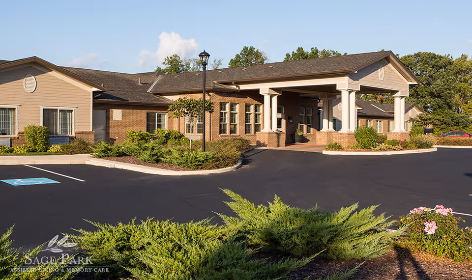 Exterior view of Sage Park Assisted Living & Memory Care facility showing a single-story building with brick and beige siding, a covered entrance supported by white columns, surrounding greenery, a parking lot with a handicapped parking space, and a clear blue sky.