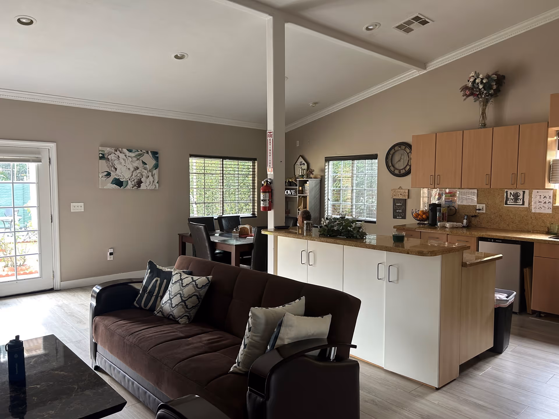 Interior view of a senior living facility showing a living room area with a brown sofa and decorative pillows, a kitchen with light wood cabinets and a granite countertop island, and a dining area with a table and chairs near windows with blinds. There is a fire extinguisher mounted on a white pillar and various decorative items on the walls and shelves.