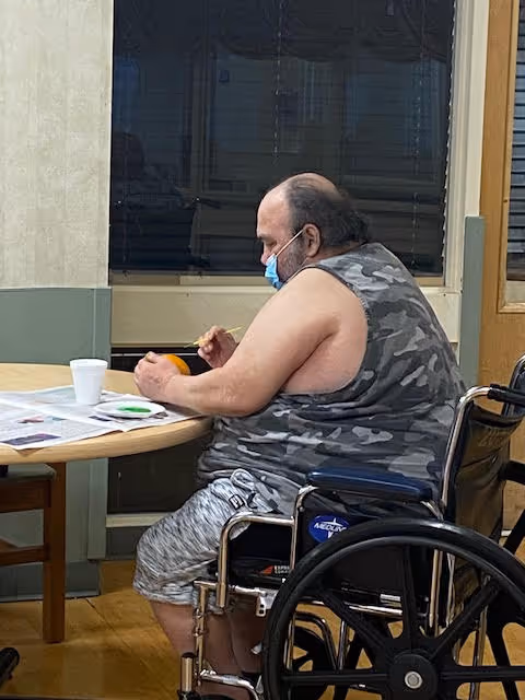 A man wearing a camouflage sleeveless shirt and shorts is sitting in a wheelchair at a round table. He is painting an orange object with a small brush. There are newspapers, a white cup, and a small paint palette on the table. The room has a window with closed blinds and light-colored walls.