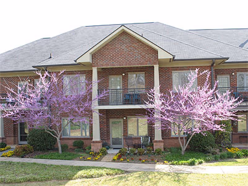 Two-story brick building facade with balconies, entry doors, and flowering pink trees and landscaped beds in front.
