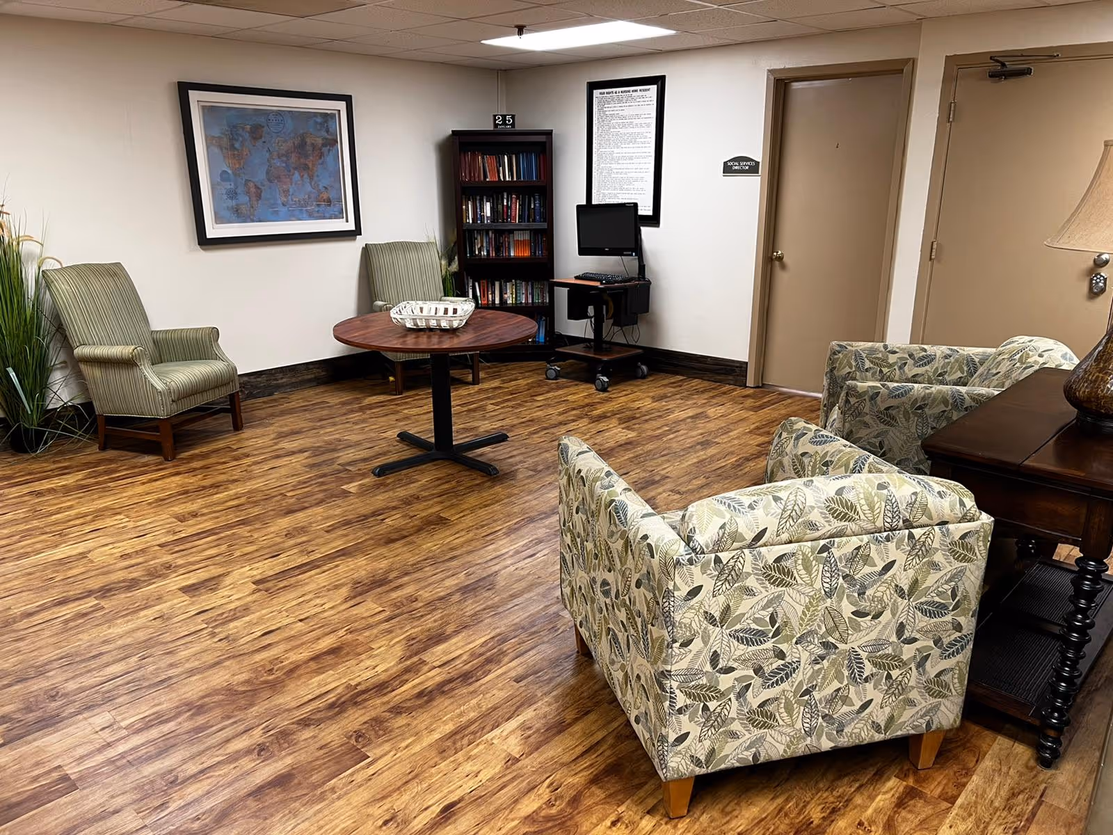 A senior living common room with patterned armchairs, a round table, bookshelf, and a small computer station against a wall.