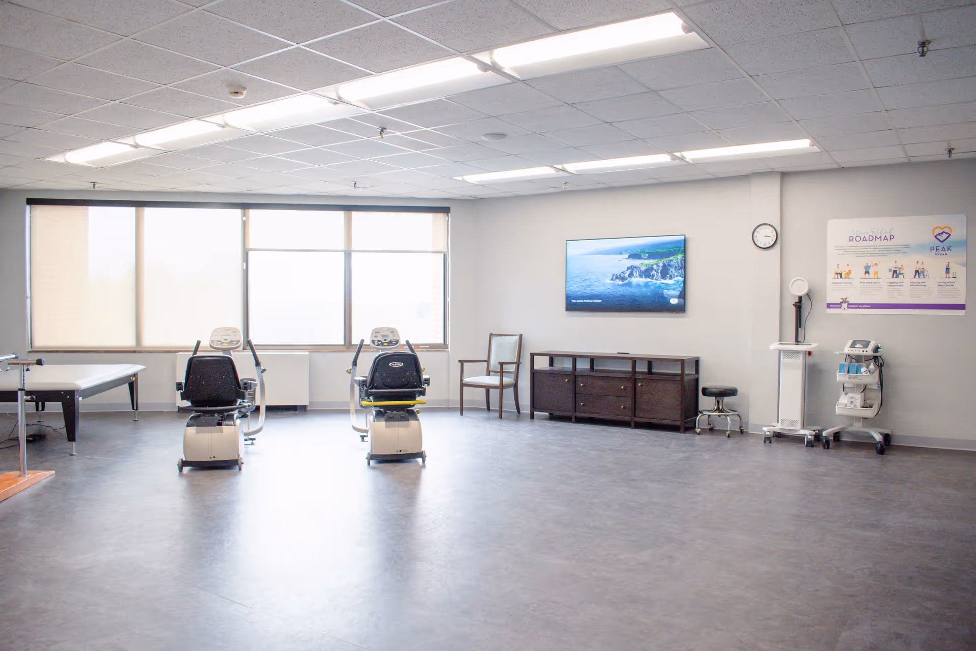 A spacious rehabilitation or therapy room with two exercise bikes facing a wall-mounted TV. The room has large windows with blinds, a treatment table on the left, a wooden chair, a dark wooden cabinet, medical equipment on carts, and a poster on the wall. The floor is smooth and the ceiling has fluorescent lighting.